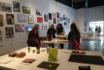 Art Exhibition, paintings on the wall tables with books and design objects, students looking at the objects